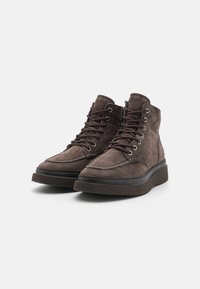 Pantofi înalți din suede maro, cu sireturi, având o talpă mai închisă la culoare și detalii cusute pe partea superioară. Suprafaț�ă texturată.