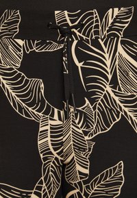 Svarta byxor med ett stort beige lövmönster, med dragsko i midjan och en slät textur. Designen betonar tropiska lövmönster.