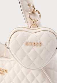 Λευκή καπιτονέ τσάντα χειρός με προσαρτημένη θήκη σε σχήμα καρδιάς. Διαθέτει χρυσά μεταλλικά στοιχεία και ανάγλυφο λογότυπο "GUESS" στο μπροστινό μέρος. Επιφάνεια με υφή.