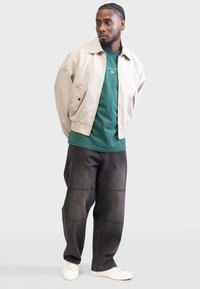 Veste beige à col portée sur une chemise verte, pantalon large noir avec des coutures en panneau, et baskets blanches, se tenant sur un arrière-plan neutre.