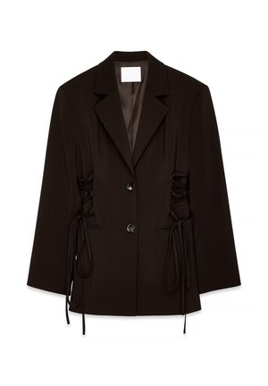 Donna che indossa un blazer marrone scuro aderente con laccetti laterali, abbottonato davanti, abbinato a una gonna marrone scuro coordinata davanti a una parete semplice.