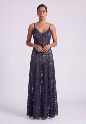 Donna in piedi che indossa un lungo vestito in pizzo blu navy trasparente con spalline sottili e biancheria intima color carne, mani leggermente intrecciate in vita.
