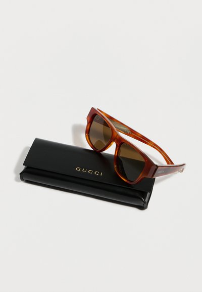 Lunettes de soleil Gucci teintées marron reposant sur un étui Gucci en cuir noir avec des lettres dorées sur un fond blanc.