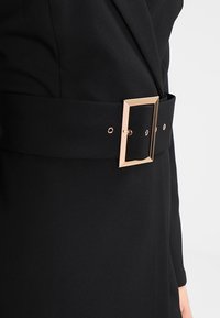 Gros plan d'une ceinture en tissu noir avec une grande boucle rectangulaire dorée portée autour de la taille sur des vêtements noirs.