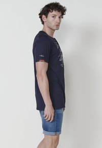 Navy zils T-krekls, kas izgatavots no kokvilnas, ar apaļu kakla izgriezumu, īsām piedurknēm ar sarkaniem akcentiem un balto grafisko uzdruku uz priekšpuses.