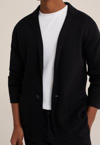 Svart cardigan med texturerad stickning, sjalkrage, en knappstängning och två framfickor. Bärs över en vit t-shirt.