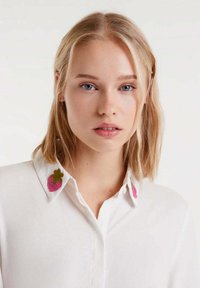 Joven mujer con cabello rubio que lleva una camisa blanca con fresas rosas bordadas en el collar, mirando hacia adelante.