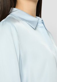 Blusa di raso azzurro chiaro con colletto classico, chiusura a bottoni e texture liscia, che mette in risalto un'elegante drappeggiatura e una silhouette slanciata.