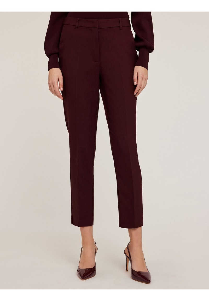Femme portant un pantalon court bordeaux sur mesure et des talons aiguilles assortis, debout devant un fond neutre.