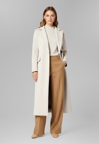 Lungo cappotto crema in tessuto testurizzato, pantaloni a gamba larga marroni e scarpe color beige chiaro. Il cappotto presenta un colletto e tasche nascoste.