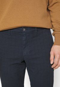 Marina-rutiga byxor med slim fit, med knappstängning och subtil textur. Kombineras med en ljusbrun långärmad skjorta.