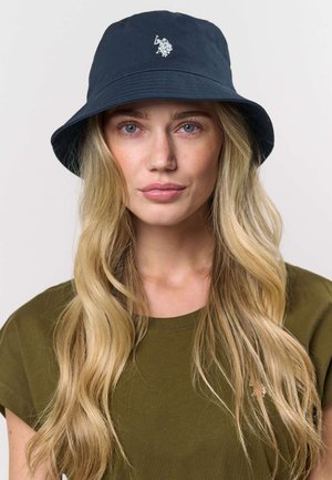 Tumši zilas auduma spainīša cepure ar gludu tekstūru un priekšpusē nelielu baltu logotipa akcentu.