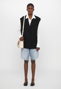 Svart oversized vest med brede revers, knappelukking, kombinert med en hvit skjorte, denimshorts og en liten beige crossover-veske.