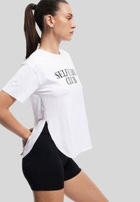 Camiseta blanca con la inscripción "SELF CARE CLUB" impresa en negro. Presenta mangas cortas, aberturas laterales y se combina con pantalones cortos negros. Textura suave.