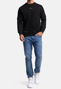Mand iført en sort sweatshirt med teksten "WEAR THE CODE", blå jeans og hvide høje sneakers, går med den ene hånd i lommen.