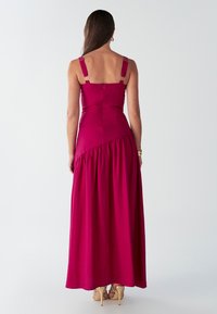 Vestido maxi ajustado en la parte posterior en un vibrante color magenta, con tirantes anchos, bajo con volantes y una textura suave y fluida. Detalles en hardware tono dorado.