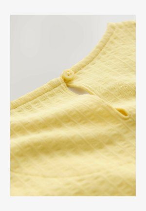 Sárga ruhadarab közeli kép, amely texturált szövetet mutat be rácsmintával, kerek gombos záródással és nyakkivágás részletekkel.