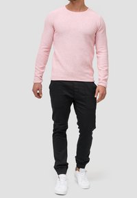 Helles rosa Strickpullover mit langen Ärmeln und Rundhalsausschnitt, kombiniert mit schwarzen Jogginghosen und weißen Sneakern. Einfaches Design und gedämpfte Farben.