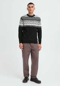 Schwarzer und weißer gestrickter Pullover mit geometrischen Mustern, kombiniert mit braunen Hosen und schwarzen Schuhen. Das Modell steht vor einem hellen Hintergrund.