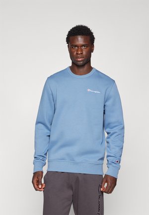 Sudadera de algodón azul con cuello redondo y puños acanalados. Presenta un pequeño logotipo rojo y blanco en el pecho. Se lleva con pantalones grises.