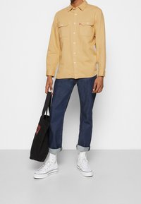 Chemise boutonnée jaune clair avec deux poches poitrine, associée à un jean bleu marine et des baskets montantes blanches. Sac cabas noir avec logo rouge.