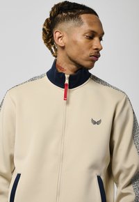 Veste zippée beige avec un col haut, des accents navy, des manches à motif texturé et un petit logo navy sur la poitrine.