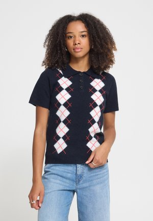Femme aux cheveux bouclés debout, portant un polo bleu marine à manches courtes avec un motif losanges blancs et rouges, assorti à un jean bleu clair.