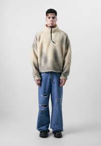 Fleece-Pullover, creme mit verlaufenden grauen Flecken, hoher Kragen und schwarzem Kordelzug; kombiniert mit distressed blauen Jeans, mit gespritzten weißen Details.