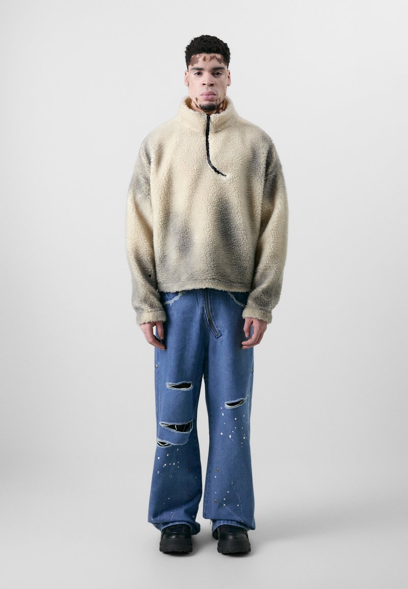 Fleece-Pullover, creme mit verlaufenden grauen Flecken, hoher Kragen und schwarzem Kordelzug; kombiniert mit distressed blauen Jeans, mit gespritzten weißen Details.