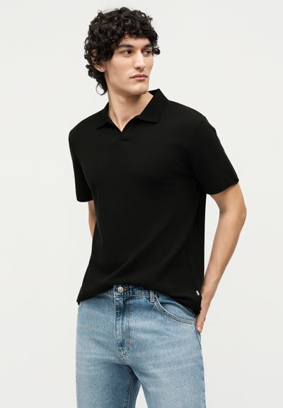 Jeune homme aux cheveux bouclés portant un polo noir à manches courtes et un jean bleu clair, les mains dans les poches, regardant sur le côté.