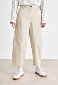 Pantaloni beige a gamba larga e cropped con tasche frontali, indossati con una camicia bianca a righe, calzini bianchi e sneakers bianche con suole color gomma.