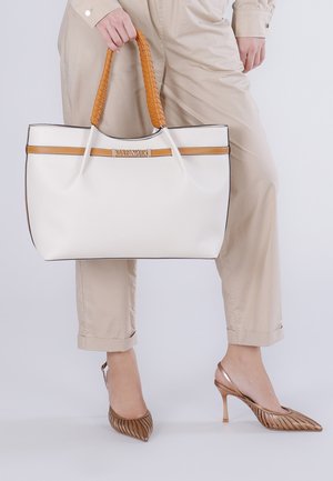 Persona che indossa pantaloni beige e tacchi bronzo che tiene una grande borsa bianca con manici intrecciati marroni e rifiniture.