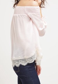 Žena v off-shoulder světle růžové blůze s krajkovým lemem, která je zkombinována s tmavě modrými džíny, zobrazená zezadu a z boku.