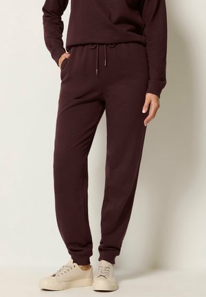 Person iført mørkebrune joggerbukser med snøreliv og sidelommer, kombineret med beige sneakers, står mod en lys baggrund.