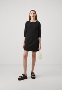 Svart klänning med kort ärm och rund halsringning, kombinerad med kraftiga svarta sandaler med nitar, och en liten vit pärlhandväska.
