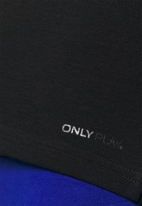T-shirt sportif noir à texture lisse et manches courtes, avec un logo argenté "ONLY PLAY" près de l'ourlet. Un tissu bleu en dessous est visible.