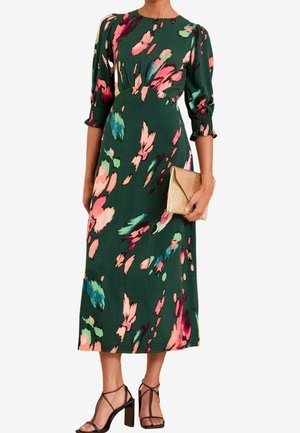 Kvinde iført en grøn midi-kjole med pink, blå og røde abstrakte blomsterprint, der holder en guldtaske og har sorte højhælede sandaler på.