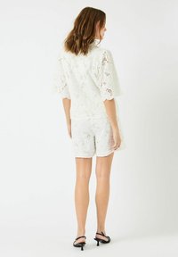 Hvitt blondertop med korte ermer kombinert med matchende blondeshorts. Har et blomsterdesign, åpen vev og rett saum. Svarte hælsandaler.