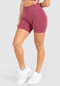 Burgundské vysoké cyklistické šortky vyrobené z elastického materiálu mají žebrovaný pas a hladký povrch. Bílé tenisky dokončují outfit.