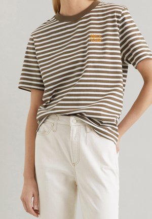 Vrouw draagt een bruin-wit gestreept shirt met het logo "Marc O'Polo Denim" en een hoge taille off-white broek, hand in de zak.