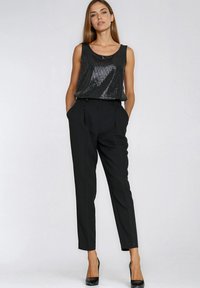 Kobieta ubrana w czarny top bez rękawów z cekinami, czarne eleganckie spodnie z wysokim stanem oraz czarne szpilki, stojąca z rękami w kieszeniach.