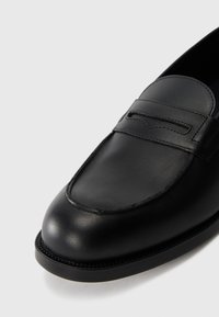 Mocasini negri din piele cu vârful rotunjit, având un design cusut și un detaliu ridicat pe partea din față. Textură netedă, accente minime.