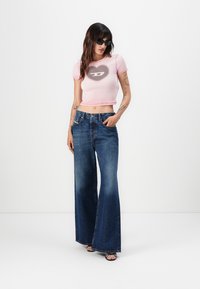 Pink cropped t-shirt featuring a heart graphic, paired with flared dark blue denim jeans. Model wearing black sunglasses and strappy sandals.