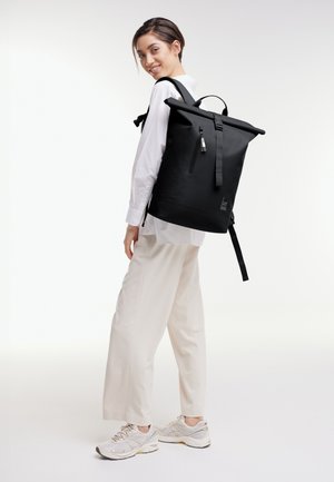 Mujer joven con camisa y pantalones blancos, llevando una gran mochila enrollable negra, sonriendo y mirando hacia atrás por encima del hombro.