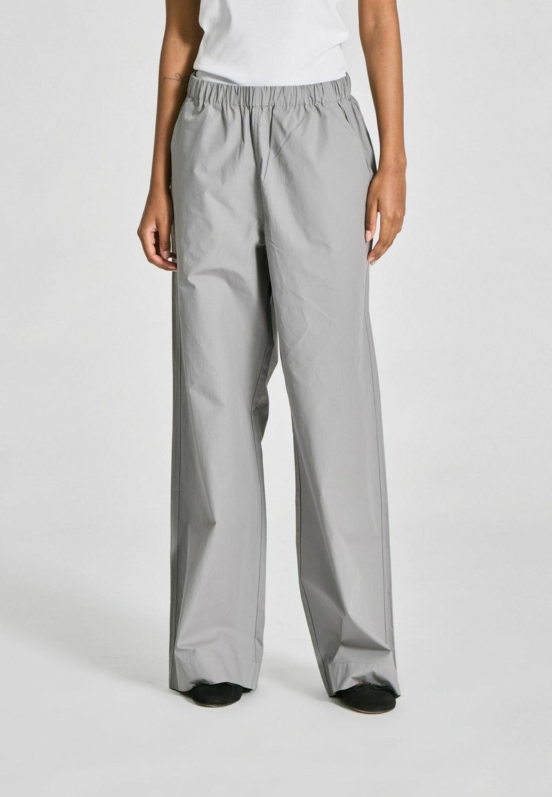 Personne portant un pantalon ample gris clair taille haute avec ceinture élastique, associé à des chaussures noires sur un fond clair uni.
