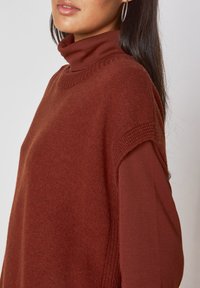 Maglione a collo alto di colore ruggine, con tessuto testurizzato, vestibilità comoda e spalle svasate. Caratterizzato da un collo a coste e maniche lisce.