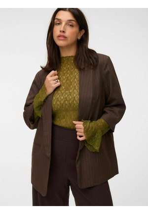 Woman wearing a brown pinstripe blazer over an olive green lace top with flared sleeves and dark brown pants, posing against a white background.