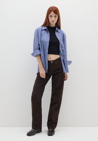 Modella che indossa una camicia a quadri blu con bottoni, un crop top nero e pantaloni maculati marroni con stivali neri a punta quadrata.
