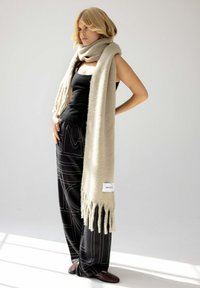 Écharpe beige en matériau doux, avec des bords frangés. Portée par-dessus un haut noir, associée à un pantalon foncé à motifs et des chaussures sombres.