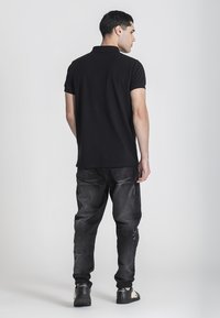 Černé polo tričko s krátkými rukávy, kombinované s roztrhanými tmavými džíny. Modelka stojí bokem a předvádí jednoduchý a ležérní outfit.